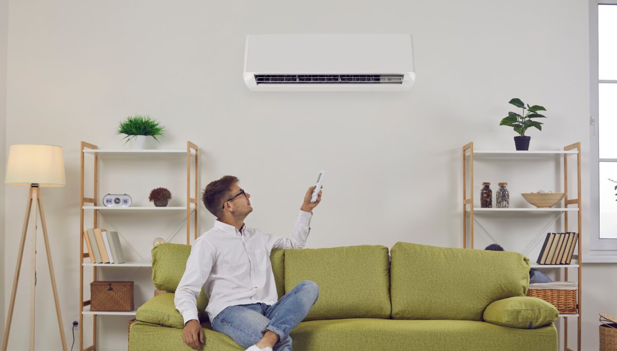 Man sitting on a green sofa in a modern living room, pointing a remote control at a wall-mounted air conditioner.
