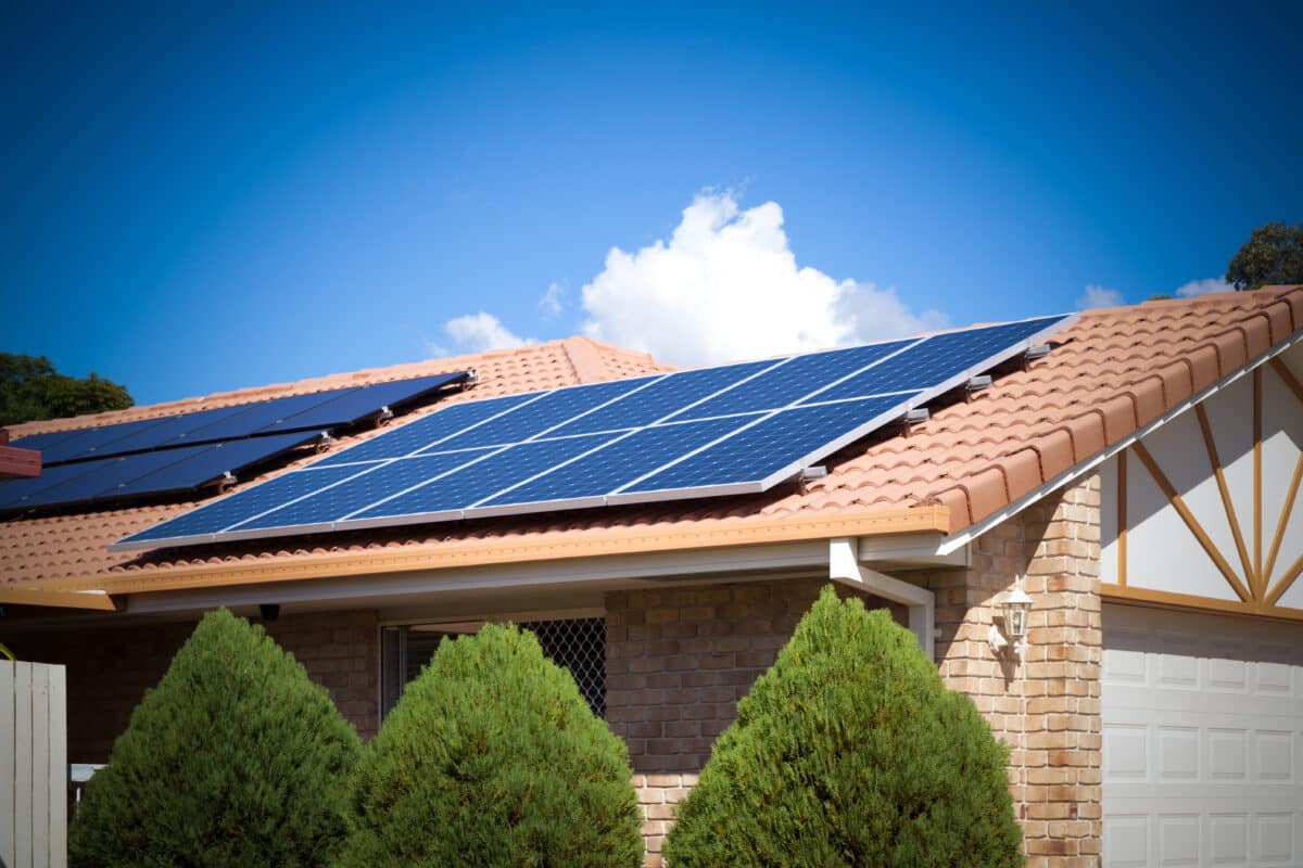 Brick house with terracotta tiled roof fitted with rooftop solar panels, set against a bright blue sky.