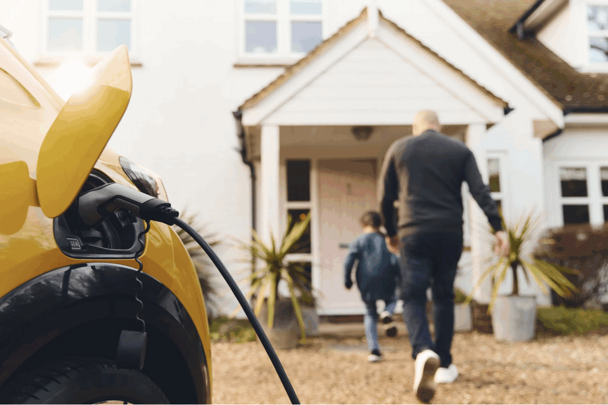 Yellow electric vehicle charging in a driveway while a man and child walk toward a white house.