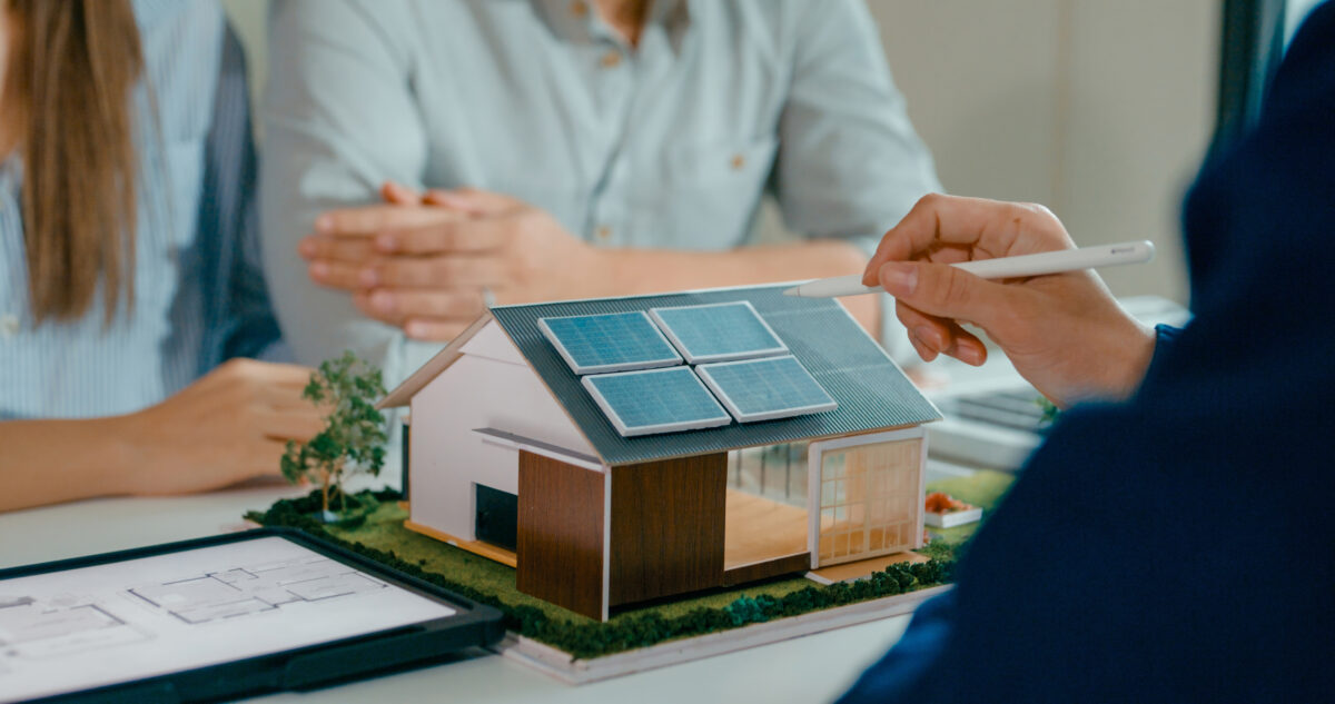Close-up of a meeting where a person is pointing to a scale model of a house with solar panels on the roof. Two other people sit across the table, and a digital tablet displaying floor plans is visible in the foreground.