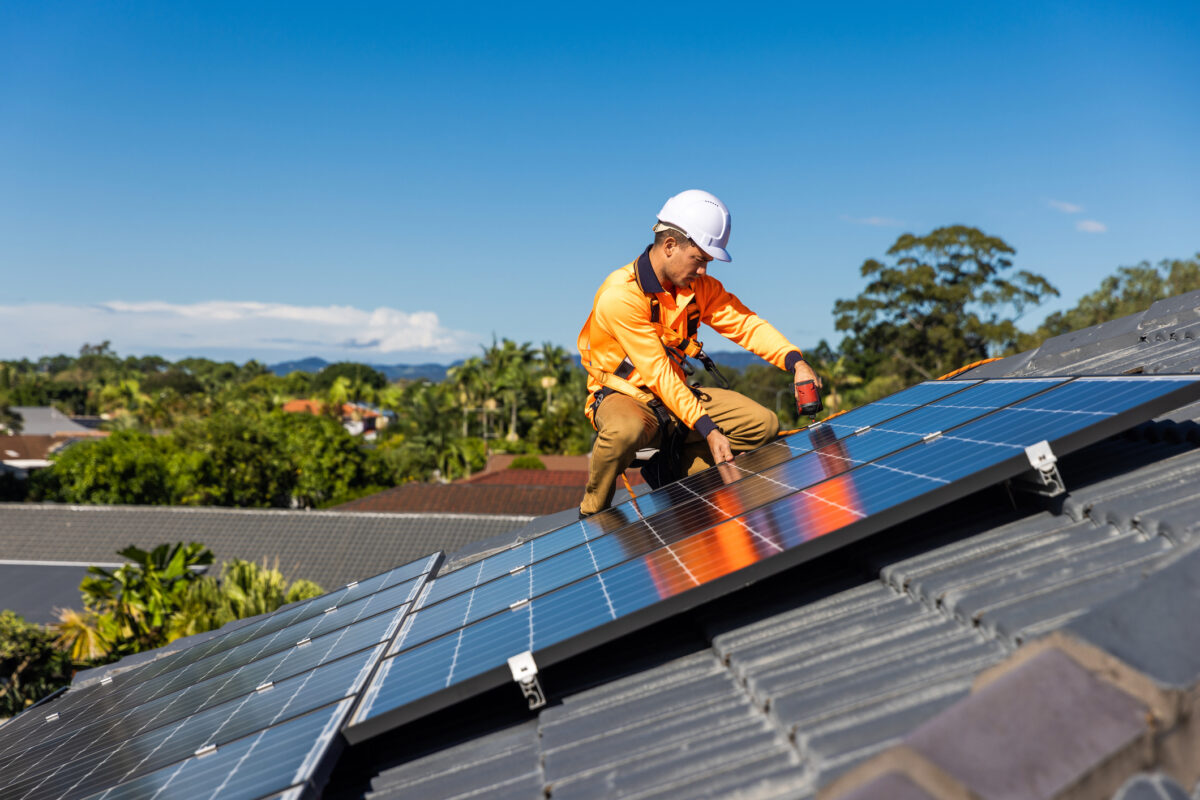 Electrician in high-visibility clothing and a safety helmet installing solar panels on a tiled residential roof under a clear blue sky, with trees and rooftops visible in the background.