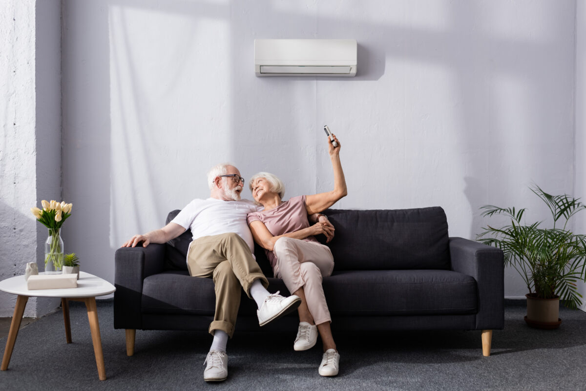 Older couple sitting on a sofa at home, smiling and leaning together while using a remote to control a wall-mounted split-system air conditioner.